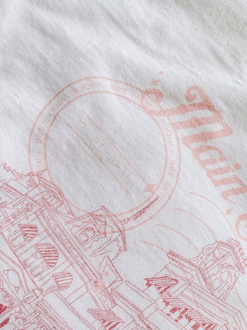 May include: A white t-shirt with a pink graphic design of a building with a circle stamp that says "Established 1982" and the text "Atlantic City" in a cursive font.