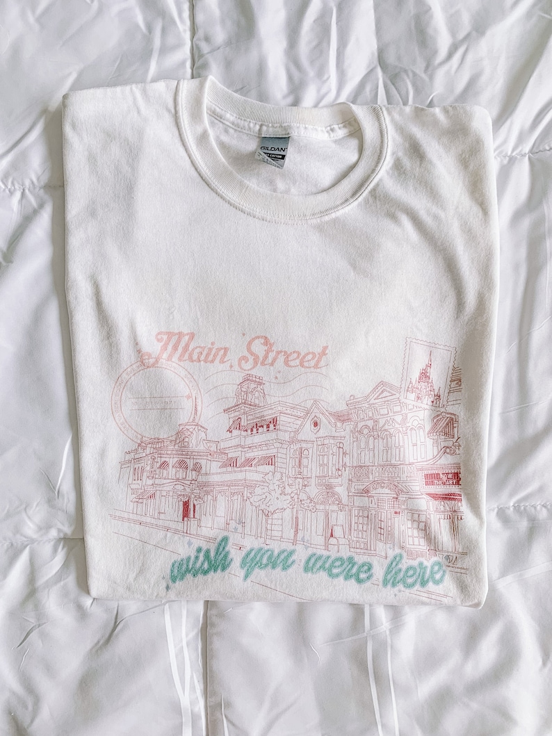 May include: White t-shirt with a pink graphic of Main Street, U.S.A. at Walt Disney World. The graphic includes the text "wish you were here".
