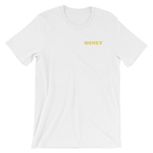 Pode incluir: Camiseta branca de manga curta com a palavra "HONEY" impressa em amarelo na frente.