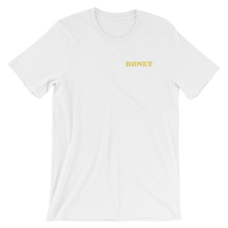 Pode incluir: Camiseta branca de manga curta com a palavra "HONEY" impressa em amarelo na frente.