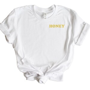 Pode incluir: Camiseta branca com um gr&aacute;fico "HONEY" dourado na frente.