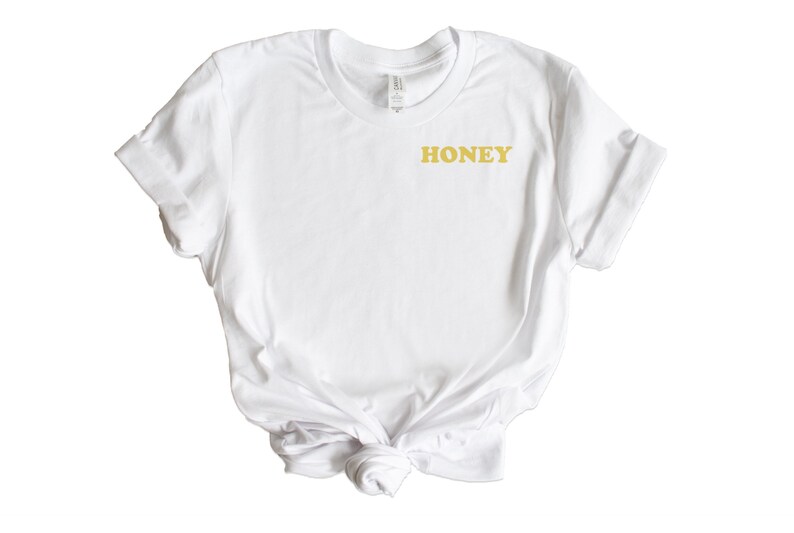Pode incluir: Camiseta branca com um gr&aacute;fico "HONEY" dourado na frente.