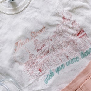 May include: White t-shirt with a pink and green graphic print of a street scene with buildings and the text "Main Street" and "wish you were here".