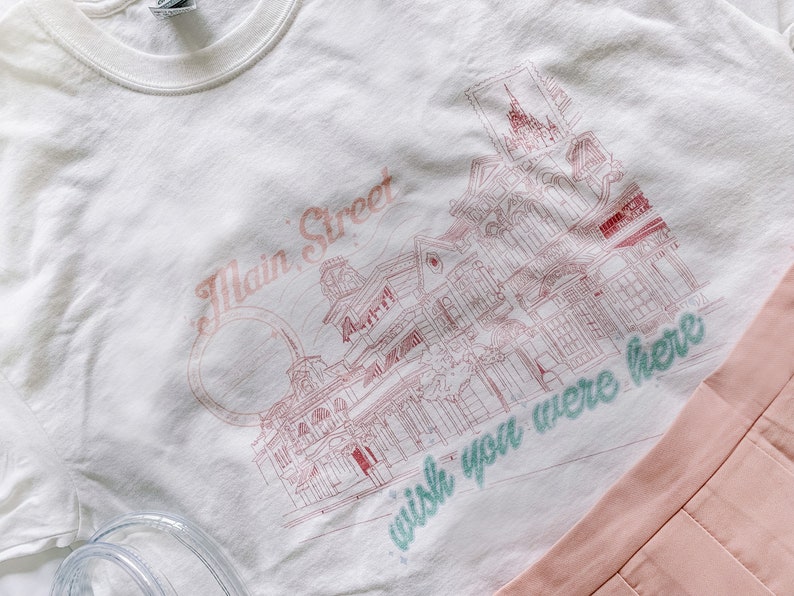 May include: White t-shirt with a pink and green graphic print of a street scene with buildings and the text "Main Street" and "wish you were here".