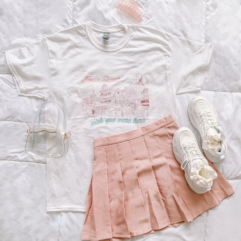 May include: White t-shirt with a graphic of Main Street, a building with the text "wish you were here". A pink pleated skirt, white sneakers, and a clear plastic bag are also pictured.
