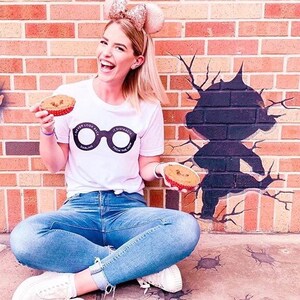 May include: A woman wearing a white t-shirt with a graphic of a pair of glasses that says "I never look back darling, it distracts from the view". She is holding two mini pies in her hands and is sitting in front of a brick wall with a black silhouette of a person painted on it.