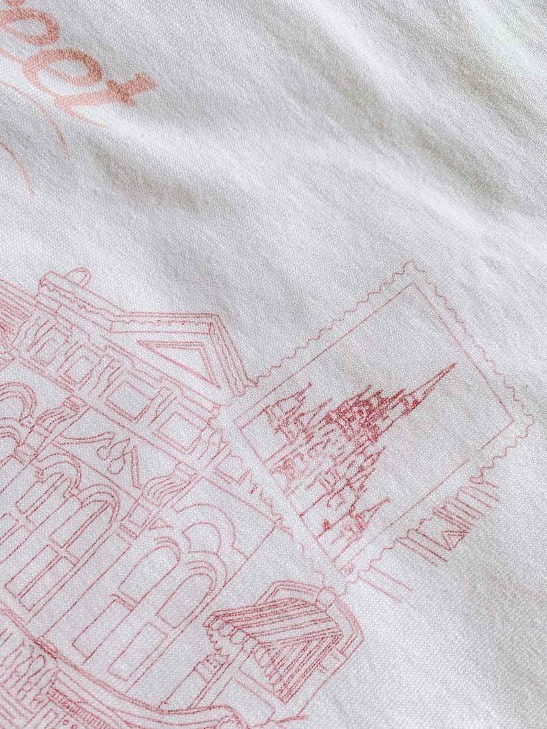 May include: A white fabric tote bag with a pink outline of a building and a stamp with a pink outline of a castle. The text "Book" is printed in pink on the bag.