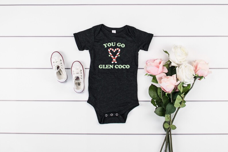 May include: A black baby onesie with the text "You go Glen Coco" and a candy cane heart graphic. The onesie is on a white wooden surface with a pair of white sneakers and a bouquet of pink and white roses.