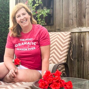 May include: A woman wearing a red t-shirt with the text "The Most Dramatic Season Ever" is sitting on a patio chair. She is holding a single red rose and there is a bouquet of red roses on a table in front of her.