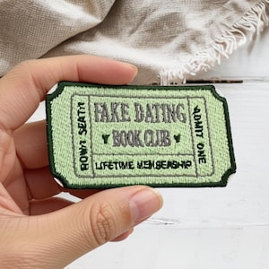 May include: A rectangular embroidered patch shaped like a vintage ticket in light green with a dark green border. The text reads FAKE DATING BOOK CLUB, ADMIT ONE, ROW 1 SEAT 1, and LIFETIME MEMBERSHIP in dark green thread with decorative stars.