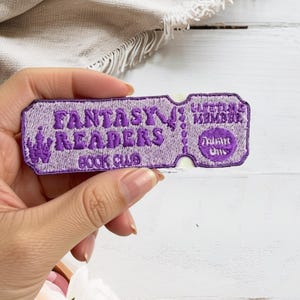 May include: A rectangular embroidered patch shaped like a ticket with purple thread on a light lavender background. It features the text FANTASY READERS BOOK CLUB, a castle icon, the words LIFETIME MEMBER, and a circular ADMIT ONE graphic.