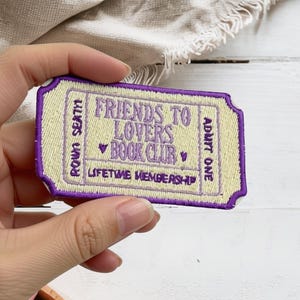 May include: A rectangular embroidered patch shaped like a ticket with a cream-colored background and a purple border. The text reads FRIENDS TO LOVERS BOOK CLUB, ROW N SEAT H, ADMIT ONE, and LIFETIME MEMBERSHIP, accented with two small purple hearts.