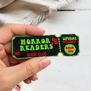 May include: A rectangular embroidered patch shaped like a ticket with a black background and bright green border. It features the text "HORROR READERS BOOK CLUB" in green, a red handprint, and the text "LIFETIME MEMBER" with a red circular graphic.