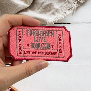 May include: A rectangular embroidered patch shaped like a ticket with a pink background and dark pink border. The text reads FORBIDDEN LOVE BOOK CLUB, ADMIT ONE, ROW 1 SEAT 1, and LIFETIME MEMBERSHIP in dark gray thread with small heart icons.