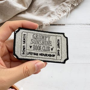 May include: A rectangular embroidered patch shaped like a vintage ticket with a black border and light gray background. It features the text GRUMPY SUNSHINE BOOK CLUB, ROW 1 SEAT 4, ADMIT ONE, and LIFETIME MEMBERSHIP in black thread.