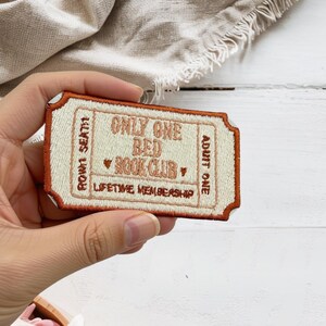May include: A rectangular embroidered patch shaped like a vintage ticket with a rust-colored border and cream-colored center. The text reads ONLY ONE BED BOOK CLUB, LIFETIME MEMBERSHIP, ROW N SEAT H, and ADULT ONE in rust-colored thread.