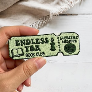 May include: A rectangular embroidered patch in light green with dark green stitching. The text reads ENDLESS TBR BOOK CLUB, LIFETIME MEMBER, and ADMIT ONE. It features an open book icon, a candle, and a ticket-style perforated edge design.