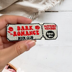 May include: A rectangular embroidered patch shaped like a ticket with a light blue background and black border. It features red text reading DARK ROMANCE BOOK CLUB, a red rose icon, and a smaller section with red text ELITE MEMBER and black text Admit One.