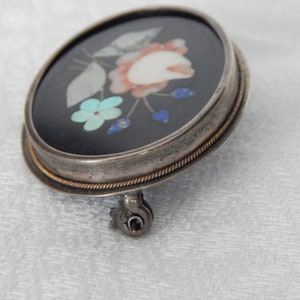 Puede incluir: Un broche de plata con fondo negro y un dise&ntilde;o floral hecho de piedras de colores. El broche tiene un pasador en la parte posterior.