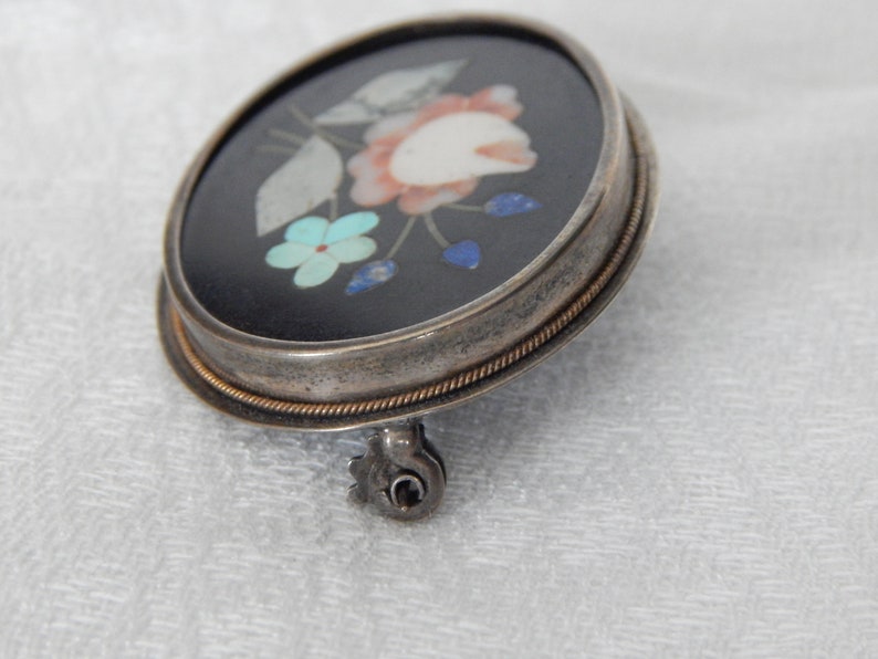 Puede incluir: Un broche de plata con fondo negro y un dise&ntilde;o floral hecho de piedras de colores. El broche tiene un pasador en la parte posterior.