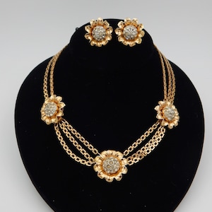 May include: A gold tone necklace and earrings set featuring a flower design with clear rhinestones. The necklace has three flower accents and a double chain.