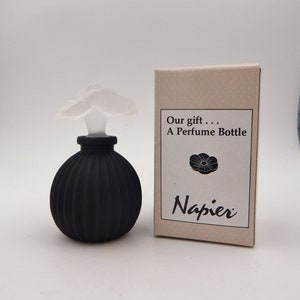 May include: A black, ribbed perfume bottle with a white, flower-shaped stopper. The bottle is next to a small box with the text "Our gift... A Perfume Bottle" and the brand name "Napier".