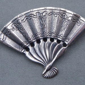 May include: A silver fan-shaped brooch with intricate floral designs and a textured surface.