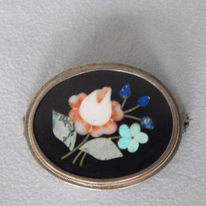 Puede incluir: Un broche de forma ovalada con un engaste de plata y un fondo negro. El broche presenta un dise&ntilde;o floral hecho de piedras de varios colores, incluyendo blanco, rosa, azul y verde.