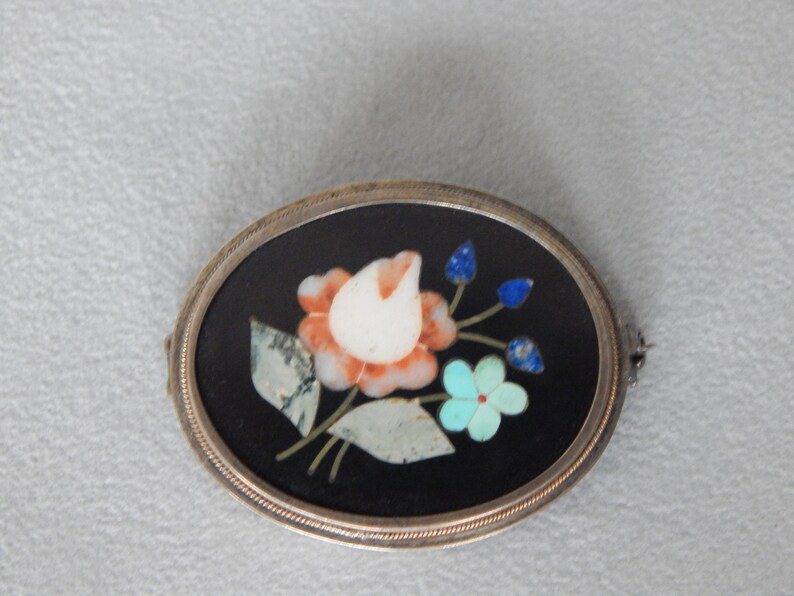 Puede incluir: Un broche de forma ovalada con un engaste de plata y un fondo negro. El broche presenta un dise&ntilde;o floral hecho de piedras de varios colores, incluyendo blanco, rosa, azul y verde.