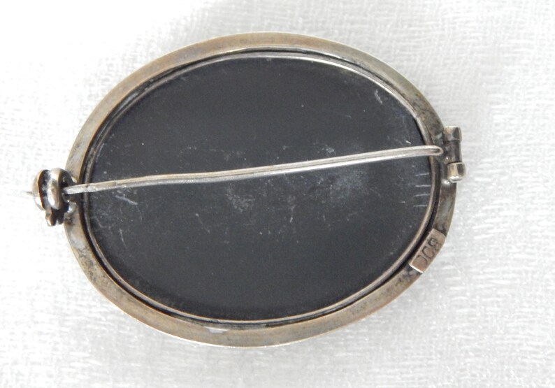 Puede incluir: Un broche ovalado de plata con un camafeo negro. La parte posterior del broche tiene un pasador de plata y un cierre. El broche est&aacute; marcado con "900" en la parte posterior.