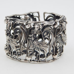 May include: A silver bracelet with an intricate design featuring two stylized birds with their wings spread. The bracelet is made of openwork metal and has a delicate, ornate look.