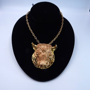 May include: A gold chain necklace with a detailed, carved, bone-like pendant of a boar's head. The pendant is set against a black velvet background.