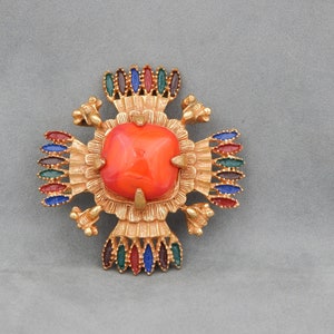 May include: A gold-toned brooch with a large, orange, faceted stone in the center. The brooch is decorated with smaller, colorful stones in a geometric pattern.