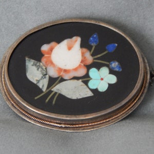 Puede incluir: Un broche ovalado de plata con fondo negro y un dise&ntilde;o floral hecho de piedras de varios colores. El dise&ntilde;o presenta una flor blanca con acentos rosados y naranjas, rodeada de hojas verdes y piedras azules y turquesas.