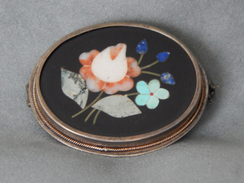 Puede incluir: Un broche ovalado de plata con fondo negro y un dise&ntilde;o floral hecho de piedras de varios colores. El dise&ntilde;o presenta una flor blanca con acentos rosados y naranjas, rodeada de hojas verdes y piedras azules y turquesas.