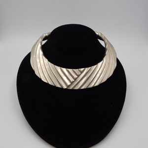 May include: A silver statement necklace with a wide, curved design. The necklace has a textured, ribbed surface.