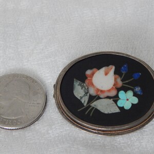 Puede incluir: Un broche ovalado de plata con fondo negro y un dise&ntilde;o floral hecho de piedras de varios colores. La flor es un tulip&aacute;n blanco con acentos rosa y naranja. Las hojas son verdes y los capullos de flores son azules.