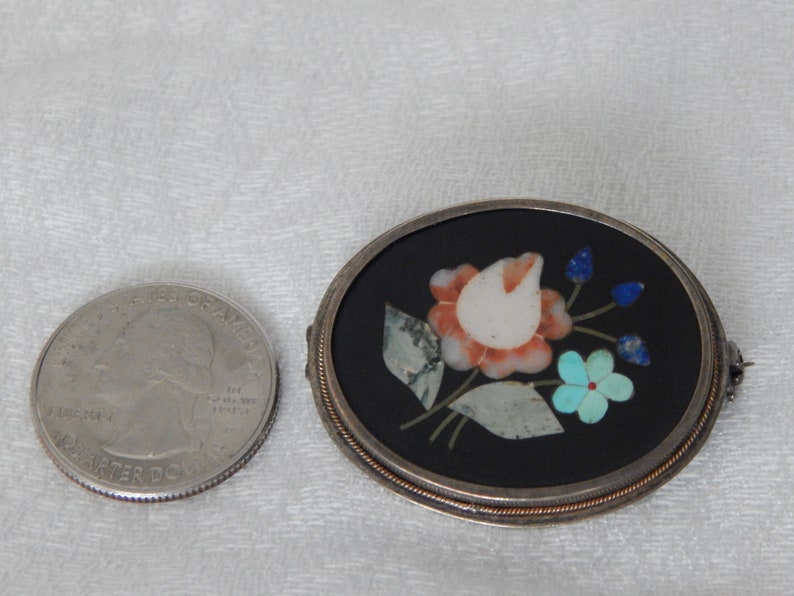 Puede incluir: Un broche ovalado de plata con fondo negro y un dise&ntilde;o floral hecho de piedras de varios colores. La flor es un tulip&aacute;n blanco con acentos rosa y naranja. Las hojas son verdes y los capullos de flores son azules.