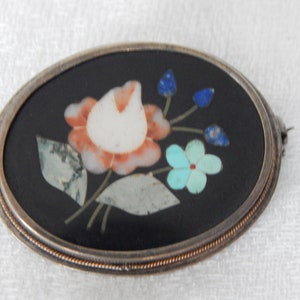 Puede incluir: Un broche de forma ovalada con fondo negro y un dise&ntilde;o floral hecho de piedras de varios colores. El dise&ntilde;o presenta una flor blanca y naranja con acentos azules y verdes.