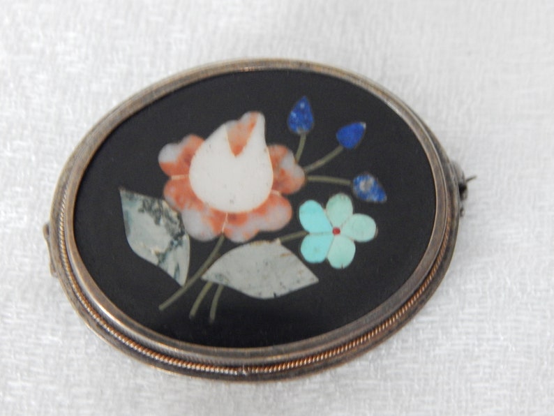 Puede incluir: Un broche de forma ovalada con fondo negro y un dise&ntilde;o floral hecho de piedras de varios colores. El dise&ntilde;o presenta una flor blanca y naranja con acentos azules y verdes.