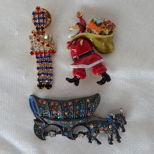 May include: Three Christmas-themed brooches featuring a red and blue jeweled soldier, a red and white Santa Claus carrying a green bag full of colorful presents, and a jeweled black and gold carriage pulled by two jeweled oxen.