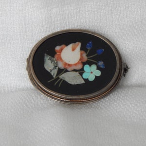 Puede incluir: Un broche redondo de plata y negro con un dise&ntilde;o floral hecho de piedras de varios colores. El broche presenta una flor blanca con p&eacute;talos rojos y naranjas, rodeada de hojas verdes y piedras azules y turquesas.