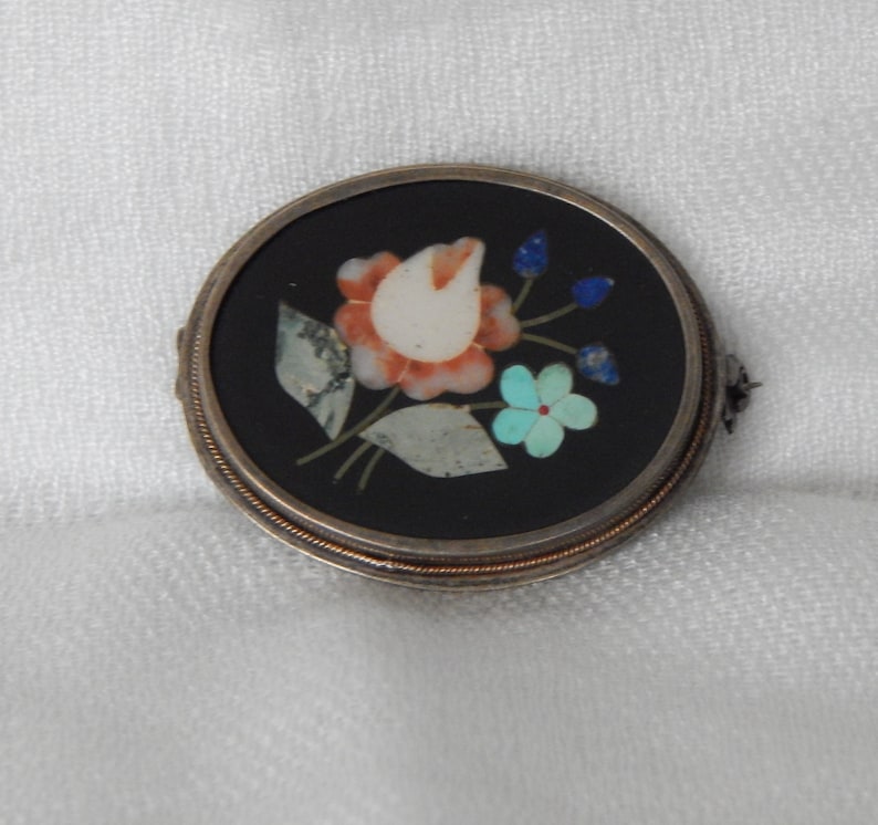Puede incluir: Un broche redondo de plata y negro con un dise&ntilde;o floral hecho de piedras de varios colores. El broche presenta una flor blanca con p&eacute;talos rojos y naranjas, rodeada de hojas verdes y piedras azules y turquesas.