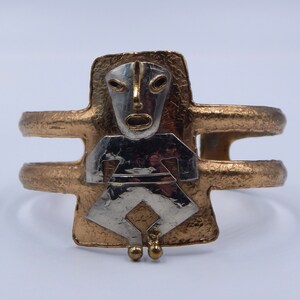 May include: A gold-toned metal bracelet with a stylized figure of a person with a face. The figure is made of silver-toned metal and is set against a textured gold background.