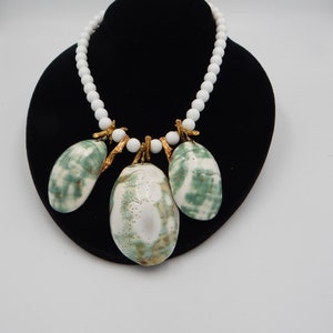 May include: A white beaded necklace with a green and white shell pendant. The pendant is made of three shells attached to the necklace with gold wire.