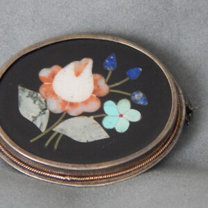 Puede incluir: Un broche ovalado de plata con fondo negro y un dise&ntilde;o floral hecho de piedras de varios colores. El dise&ntilde;o presenta una flor blanca y naranja con hojas verdes y flores azules y turquesas.