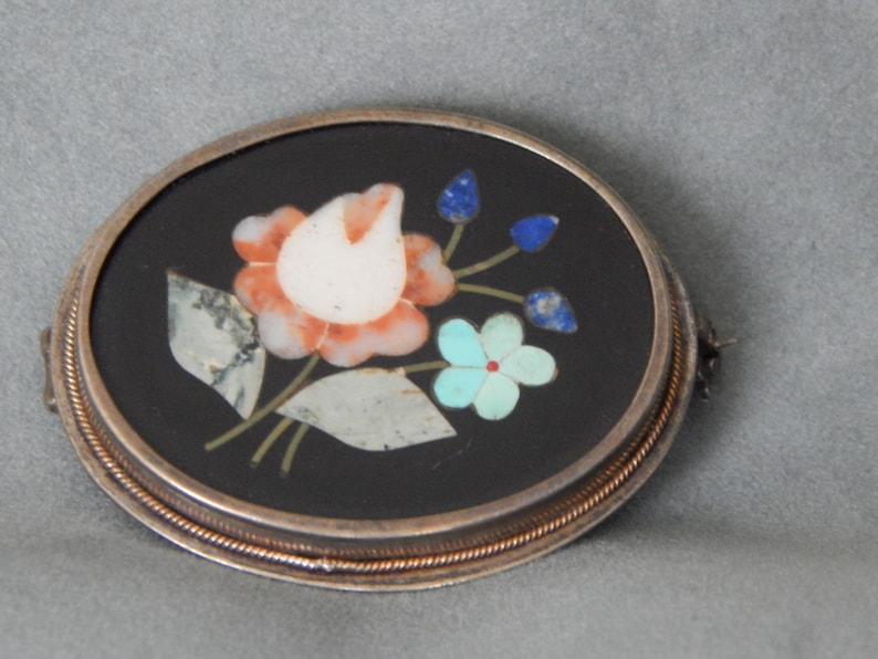 Puede incluir: Un broche ovalado de plata con fondo negro y un dise&ntilde;o floral hecho de piedras de varios colores. El dise&ntilde;o presenta una flor blanca y naranja con hojas verdes y flores azules y turquesas.