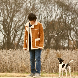 Peut inclure: Une personne portant une veste en daim marron avec un col en fourrure blanche se tient dans un champ d'herbe s&egrave;che avec un petit chien noir et blanc qui la regarde.