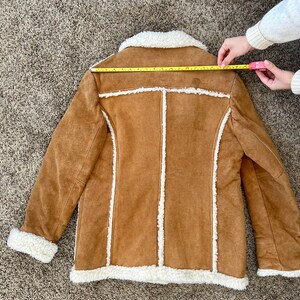Peut inclure: Une veste en daim marron avec une doublure en sherpa blanche. La veste a un col et des manches longues. La veste est pos&eacute;e &agrave; plat sur un tapis marron.