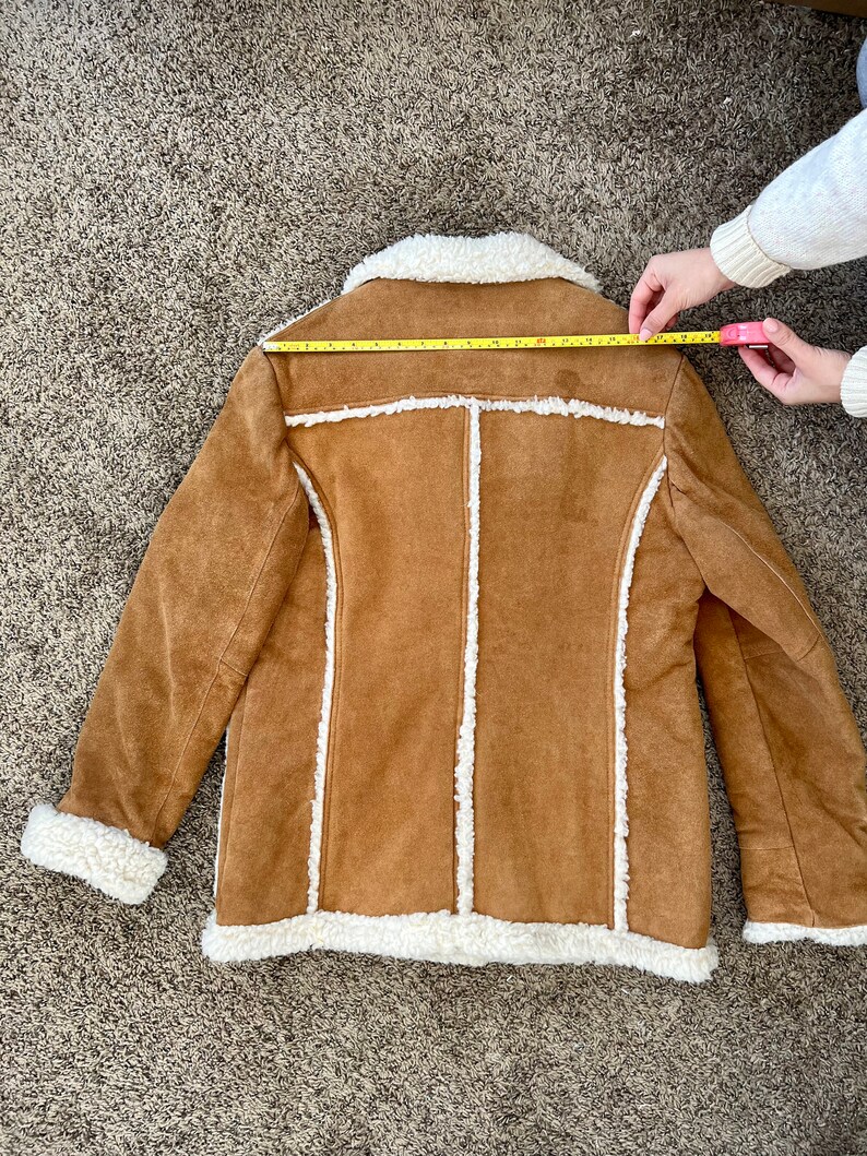 Peut inclure: Une veste en daim marron avec une doublure en sherpa blanche. La veste a un col et des manches longues. La veste est pos&eacute;e &agrave; plat sur un tapis marron.